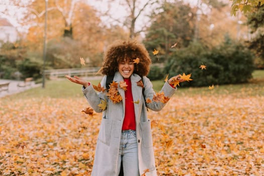 A cheerful individual with curly hair plays in a park during autumn, joyfully tossing vibrant leaves into the air among a golden blanket of fallen foliage.
