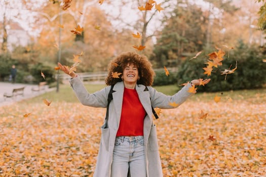 A cheerful individual with curly hair plays in a park during autumn, joyfully tossing vibrant leaves into the air among a golden blanket of fallen foliage.