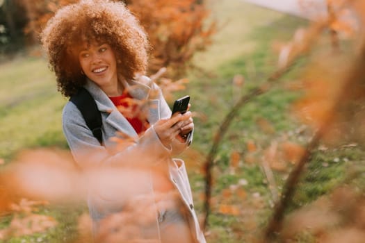 A joyful person with curly hair smiles while holding a camera, surrounded by vibrant autumn leaves in a serene outdoor setting on a sunny day.