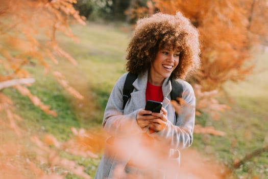 A joyful person with curly hair smiles while holding a camera, surrounded by vibrant autumn leaves in a serene outdoor setting on a sunny day.