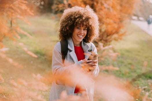 A joyful person with curly hair smiles while holding a camera, surrounded by vibrant autumn leaves in a serene outdoor setting on a sunny day.