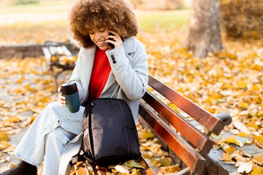 Sitting comfortably on a bench, a cheerful young woman with curly hair talks on her phone while enjoying a cup of coffee amidst fallen autumn leaves in the park.