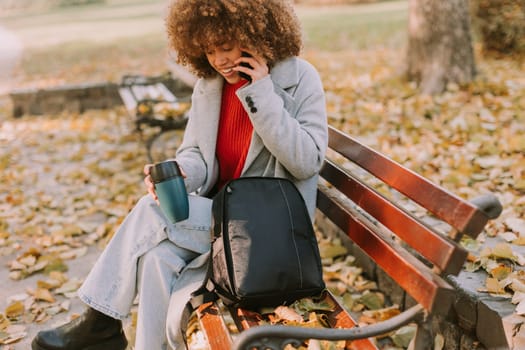 Sitting comfortably on a bench, a cheerful young woman with curly hair talks on her phone while enjoying a cup of coffee amidst fallen autumn leaves in the park.