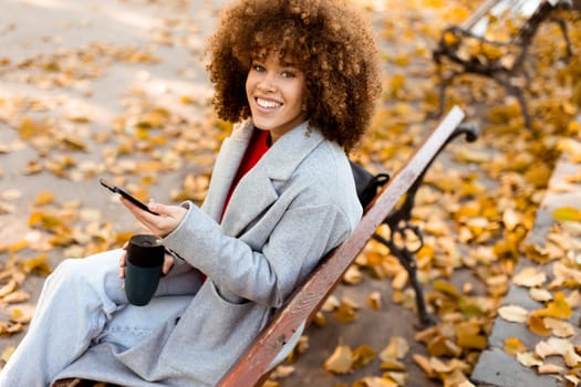 Sitting on a wooden bench in a park, a young woman smiles while texting on her smartphone, enjoying the vibrant autumn leaves that blanket the ground.