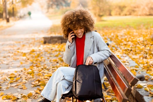 A joyful woman with curly hair makes a phone call while sitting on a park bench surrounded by vibrant autumn foliage, enjoying a beautiful day outdoors.