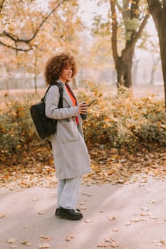 Joyful woman stands amidst vibrant autumn foliage in a park, sipping a warm drink and wearing a cozy outfit, embodying the beauty of the season in the afternoon light.