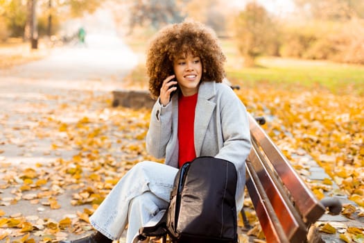 A joyful woman with curly hair makes a phone call while sitting on a park bench surrounded by vibrant autumn foliage, enjoying a beautiful day outdoors.