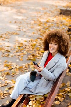 Sitting on a wooden bench in a park, a young woman smiles while texting on her smartphone, enjoying the vibrant autumn leaves that blanket the ground.