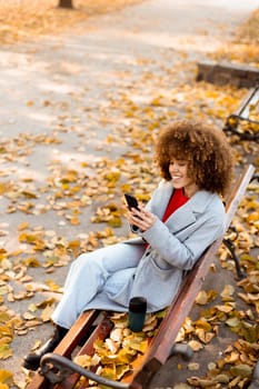 Sitting on a wooden bench in a park, a young woman smiles while texting on her smartphone, enjoying the vibrant autumn leaves that blanket the ground.