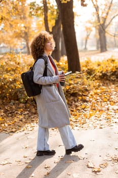 Joyful woman stands amidst vibrant autumn foliage in a park, sipping a warm drink and wearing a cozy outfit, embodying the beauty of the season in the afternoon light.