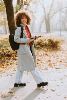 Joyful woman stands amidst vibrant autumn foliage in a park, sipping a warm drink and wearing a cozy outfit, embodying the beauty of the season in the afternoon light.