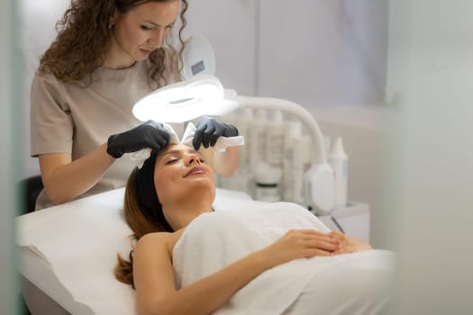 A woman enjoys a rejuvenating facial treatment at a tranquil spa. A skilled aesthetician gently wipes her forehead, enhancing her pampering experience.