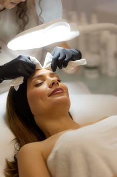 A woman enjoys a rejuvenating facial treatment at a tranquil spa. A skilled aesthetician gently wipes her forehead, enhancing her pampering experience.