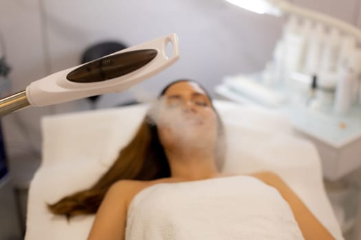 A tranquil spa setting features a woman enjoying a facial treatment, with mist enveloping her face. The atmosphere promotes relaxation and skincare rejuvenation.