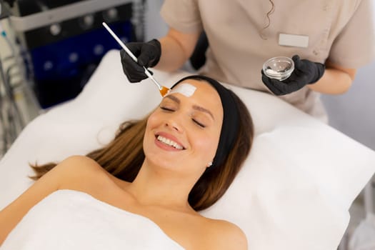 A woman lies relaxed on a treatment table in a spa while a specialist applies a soothing facial mask. Soft lighting enhances the tranquil atmosphere of self-care.