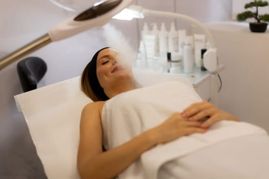 A woman comfortably reclines on a treatment bed, enjoying a calming facial steam at a serene spa. Soft light enhances the peaceful, rejuvenating atmosphere.