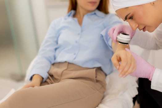 A woman sits comfortably in a modern clinic while a practitioner applies a dermatoscope device to her arm, showcasing a blend of care and advanced technology for beauty enhancement.