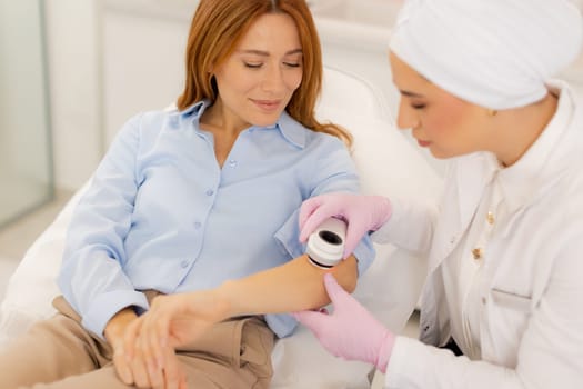 A woman sits comfortably in a modern clinic while a practitioner applies a dermatoscope device to her arm, showcasing a blend of care and advanced technology for beauty enhancement.