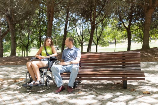 Young woman in wheelchair talking with a man sitting next to her on a park bench