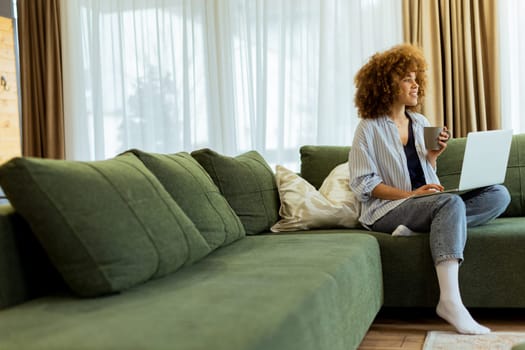 Sitting comfortably on a plush sofa, a young woman savors her warm coffee while working on her laptop, illuminated by soft natural light in a stylish living space