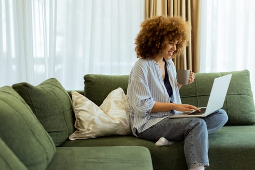 Sitting comfortably on a plush sofa, a young woman savors her warm coffee while working on her laptop, illuminated by soft natural light in a stylish living space