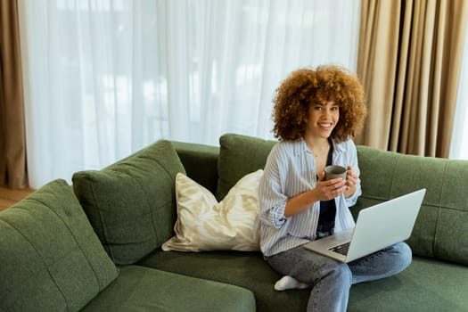 Sitting comfortably on a plush sofa, a young woman savors her warm coffee while working on her laptop, illuminated by soft natural light in a stylish living space
