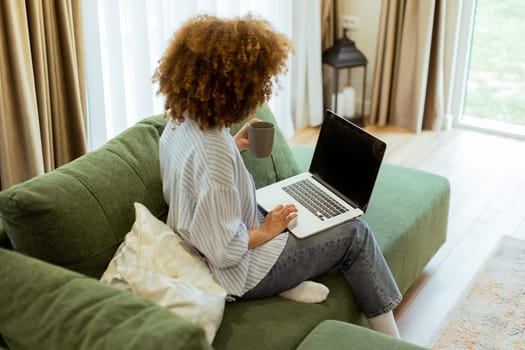 Sitting comfortably on a plush sofa, a young woman savors her warm coffee while working on her laptop, illuminated by soft natural light in a stylish living space