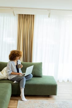 Sitting comfortably on a plush sofa, a young woman savors her warm coffee while working on her laptop, illuminated by soft natural light in a stylish living space