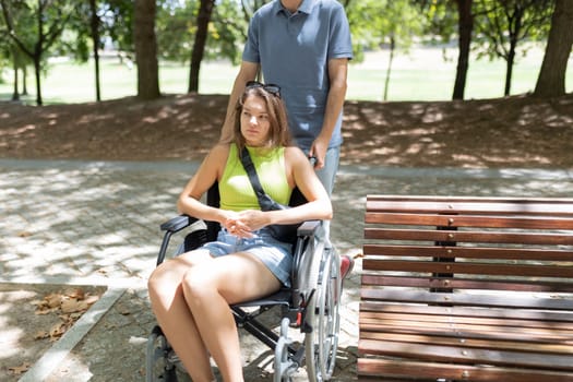 Young man pushing thoughtful disabled woman in wheelchair in park on summer day