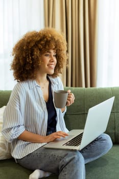 Sitting comfortably on a plush sofa, a young woman savors her warm coffee while working on her laptop, illuminated by soft natural light in a stylish living space