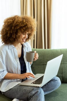 Sitting comfortably on a plush sofa, a young woman savors her warm coffee while working on her laptop, illuminated by soft natural light in a stylish living space