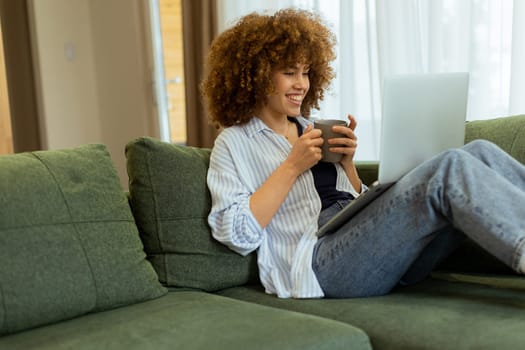Sitting comfortably on a plush sofa, a young woman savors her warm coffee while working on her laptop, illuminated by soft natural light in a stylish living space