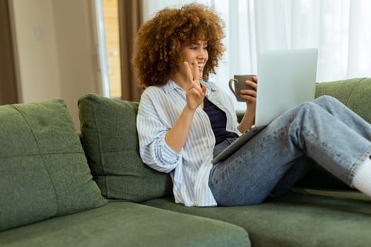Sitting comfortably on a plush sofa, a young woman savors her warm coffee while working on her laptop, illuminated by soft natural light in a stylish living space