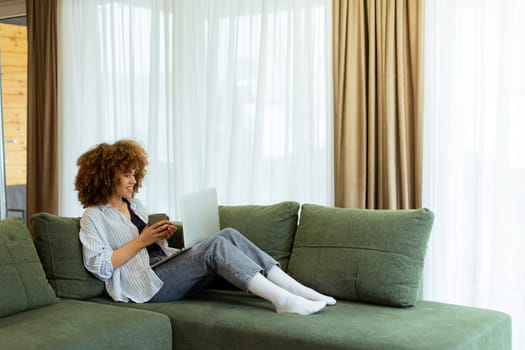 Sitting comfortably on a plush sofa, a young woman savors her warm coffee while working on her laptop, illuminated by soft natural light in a stylish living space