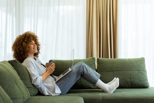 Sitting comfortably on a plush sofa, a young woman savors her warm coffee while working on her laptop, illuminated by soft natural light in a stylish living space