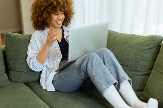Sitting comfortably on a plush green sofa, a young woman types on her laptop, basking in the warm sunlight streaming through the large windows, radiating focus and tranquility.