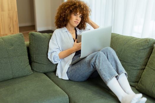 Sitting comfortably on a plush sofa, a young woman savors her warm coffee while working on her laptop, illuminated by soft natural light in a stylish living space