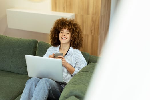 A young woman with curly hair smiles while sitting comfortably on a green sofa, using her laptop and smartphone in a bright, modern living room that exudes warmth and comfort.