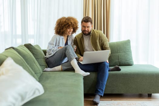 A cheerful couple sits side by side on a plush green couch, engaged in an exciting conversation as they browse through ideas on a laptop under warm sunlight streaming in