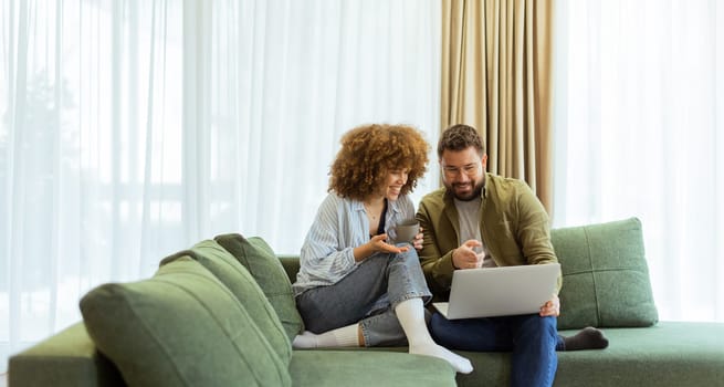A cheerful couple sits side by side on a plush green couch, engaged in an exciting conversation as they browse through ideas on a laptop under warm sunlight streaming in