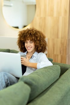 In a bright, modern living area, a young woman with curly hair smiles while using her laptop, surrounded by soft green cushions. Sunlight filters in, creating a warm atmosphere.