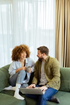 A cheerful couple sits side by side on a plush green couch, engaged in an exciting conversation as they browse through ideas on a laptop under warm sunlight streaming in