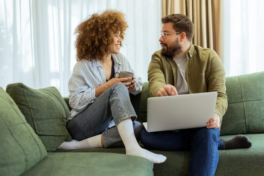 Friends share a joyful moment on a green sofa, discussing ideas while one enjoys a warm drink and the other works on a laptop, surrounded by soft natural light.