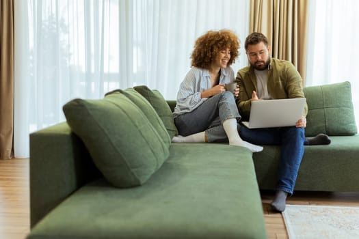 Friends share a joyful moment on a green sofa, discussing ideas while one enjoys a warm drink and the other works on a laptop, surrounded by soft natural light.