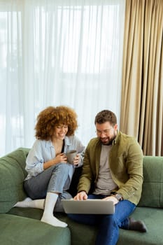 A cheerful couple sits side by side on a plush green couch, engaged in an exciting conversation as they browse through ideas on a laptop under warm sunlight streaming in