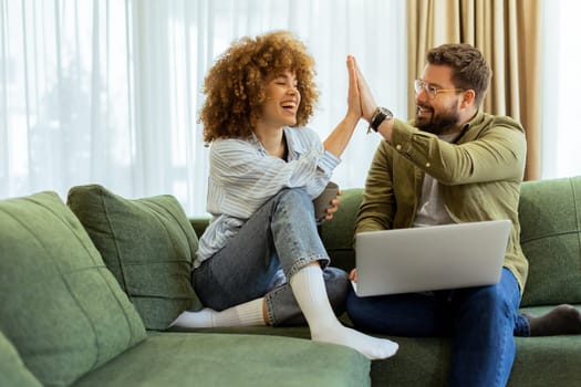 Two friends sit on a comfortable green sofa, sharing a joyful high-five while collaborating on a laptop in a bright and inviting living room.
