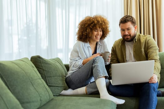 A cheerful couple sits side by side on a plush green couch, engaged in an exciting conversation as they browse through ideas on a laptop under warm sunlight streaming in