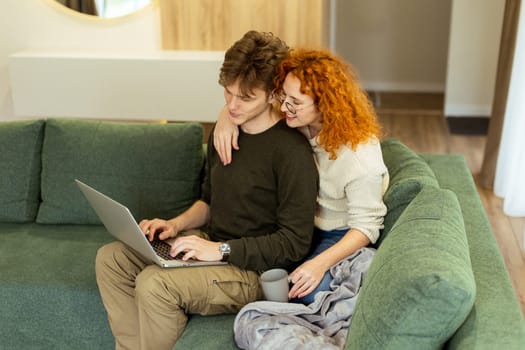 In a stylish living room, a couple shares a warm moment as they sit on a green couch, working on a laptop. The sunlight bathes them, perfect for a relaxed afternoon