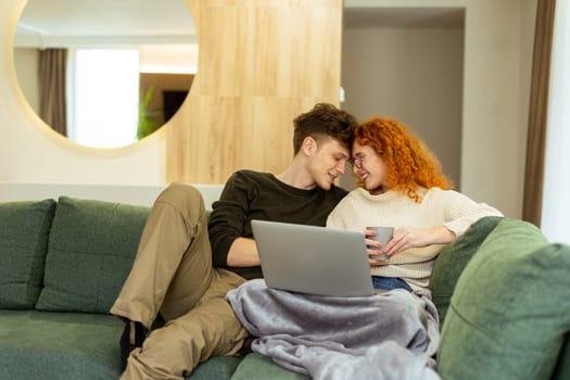 Wrapped in a soft blanket, a couple sits snugly on a green couch, sharing laughter and warmth as they browse on a laptop, sipping from a cozy mug, making delightful memories.