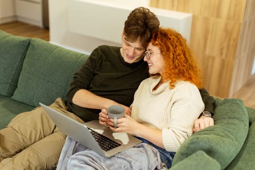 Wrapped in a soft blanket, a couple sits snugly on a green couch, sharing laughter and warmth as they browse on a laptop, sipping from a cozy mug, making delightful memories.